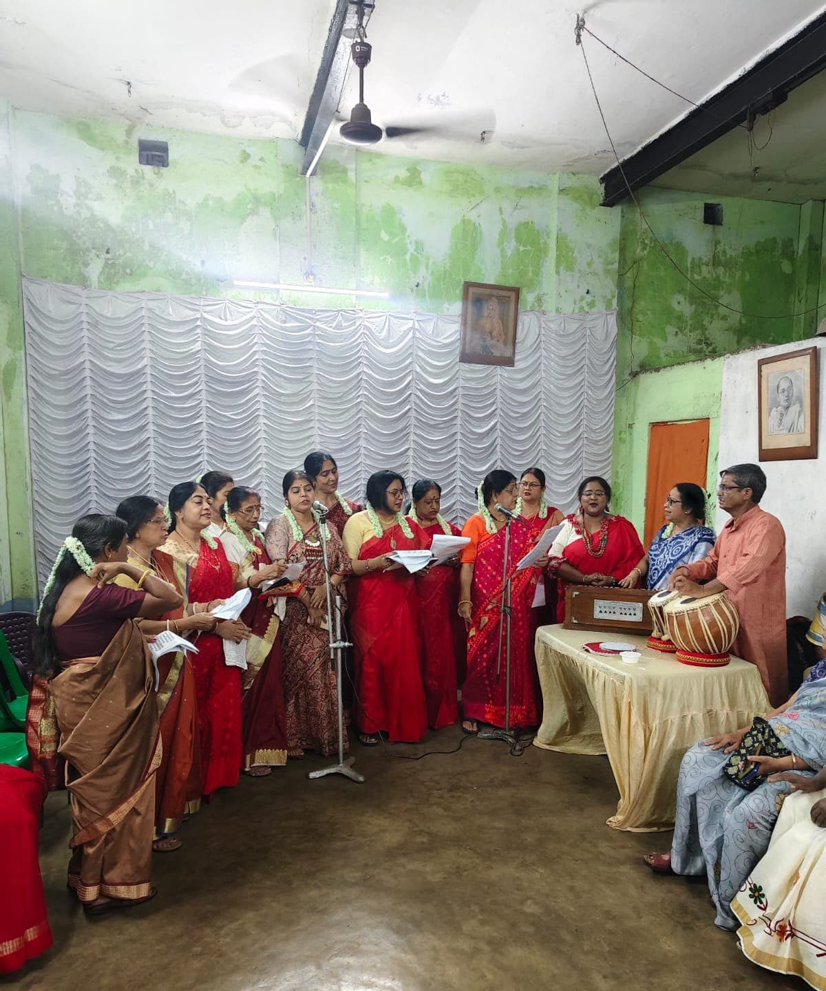 Women of the neighbourhood singing together in the club hall, accompanied on harmonium and tabla